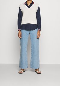 Överdimensionerad vit stickad väst över en marinblå skjorta med färggad krage, tillsammans med ljusblå vida jeans och vita sandaler.