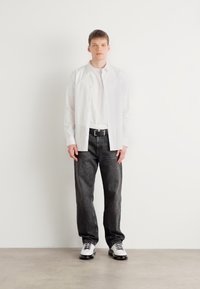 White button-up shirt over a white t-shirt, paired with dark gray jeans and black and white lace-up shoes. Simple, modern design.