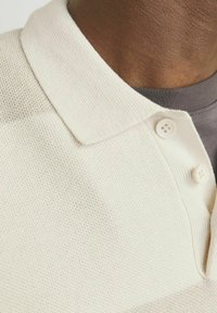 Beige polo-shirt med en struktureret stof, en krave og en lukning med tre knapper, lagt over en mørkegrå t-shirt.
