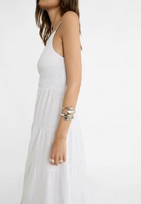 Frau in weißem ärmellosem Kleid, die ein grobes silbernes Armband am Oberarm und zwei silberne Ringe an den Fingern trägt, von der Seite gezeigt.