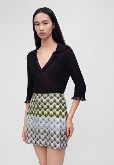 Femme aux longs cheveux noirs portant un haut noir transparent à col en V et une jupe à motif chevron vert, bleu et gris.