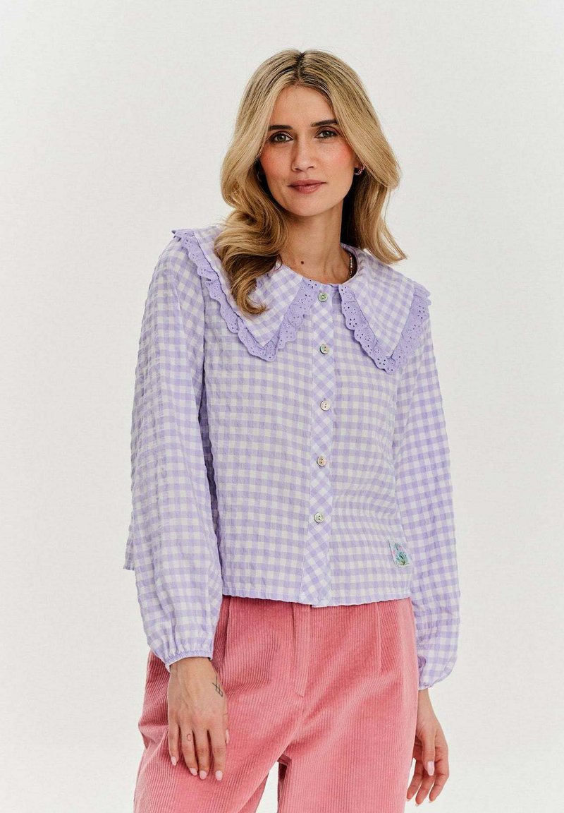 Liliowa bluzka w kratę z falbankowym kołnierzykiem, zapięciem na guziki, długimi rękawami i małym detalem na dole. W połączeniu z różowymi spodniami z corduroy.