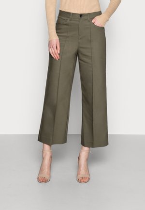 Vrouw draagt olijfgroene wijde cropped broek met een vouw aan de voorkant, beige open teen enkellaarsjes met bandje en een beige geribbelde trui.