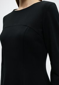 Μαύρο φόρεμα με εφαρμοστή σχεδίαση, με λαιμόκοψη bateau και διακριτικές ραφές. Κατασκευασμένο από λείο ύφασμα με δομημένη σιλουέτα.