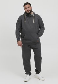 Sudadera gris con capucha y cordones, con un bolsillo frontal, combinada con pantalones de chándal grises a juego y zapatillas blancas, exhibida sobre un fondo blanco.