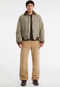 Olijfgroene bomberjack met een kraag van bruine nepbont, gecombineerd met lichtbruine losse broek met meerdere zakken en bruine schoenen.
