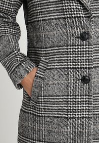 Hand i fickan på en svart och vit houndstooth-mönstrad kappa med två synliga svarta knappar.