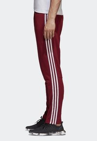 Maroon athletic pants with three white stripes running down the sides. Made of a smooth fabric, tapered at the ankles, paired with black sneakers.