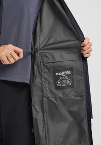 Wasserdichte Jacke aus dunkelgrauem Texapore-Material. Verfügt über einen Reißverschluss vorne, einen verstellbaren Kordelzug im Inneren und ein Pflegeetikett in der Innentasche.