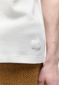 Κοντινό πλάνο ενός ατόμου που φοράει λευκό πουκάμισο με υφή και καφέ παντελόνι με υφή, εμφανίζοντας μια μικρή υφασμάτινη ετικέτα στο τελείωμα του πουκαμίσου.