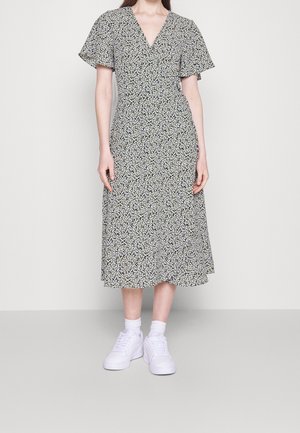 Kvinde iført en sort midi-kjole med små hvide blomster, korte flagrende ærmer, hvide sokker og hvide sneakers på en ensfarvet baggrund.