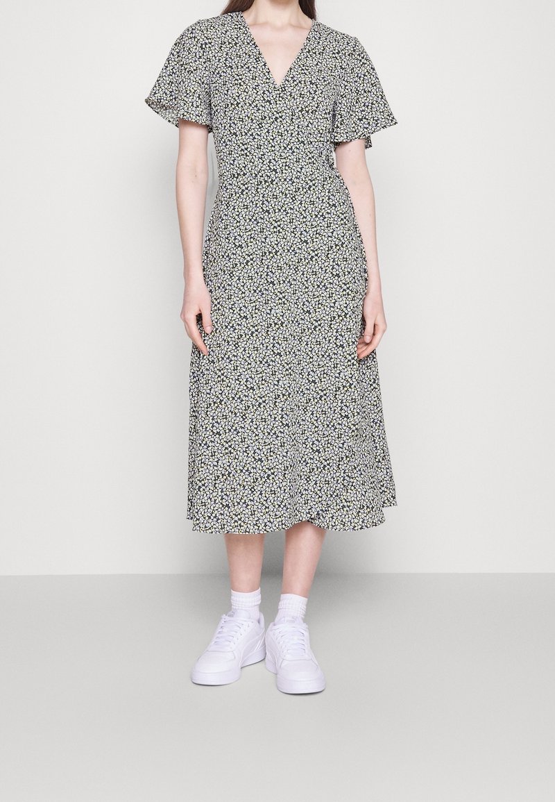Femme portant une robe midi noire avec un petit motif floral blanc, des manches courtes à volants, des chaussettes blanches et des baskets blanches sur un fond uni.