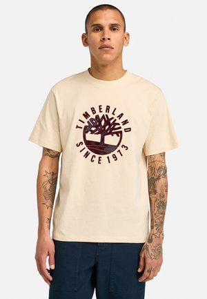 T-shirt in cotone beige con logo Timberland rosso scuro e disegno di un albero stampato sul fronte. La maglietta ha un collo rotondo classico e maniche corte.