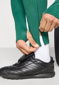 Πράσινο αθλητικό κολάν με υφασμένο μοτίβο, εφαρμοστό γύρω από τα πόδια, συνδυασμένο με μαύρα αθλητικά παπούτσια με υφασμένες λεπτομέρειες και λευκή κάλτσα.