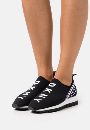 Jambes portant des baskets noires à enfiler avec le logo blanc "DKNY" et un détail à rayures sur le talon, debout sur une surface blanche.
