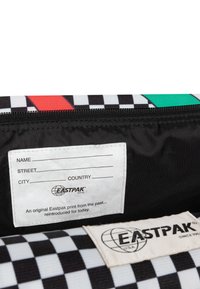 Wnętrze torby z tkaniny w czarno-białą kratkę, z białą metką zawierającą szablon adresu oraz logo Eastpak, otoczone czarną podszewką.