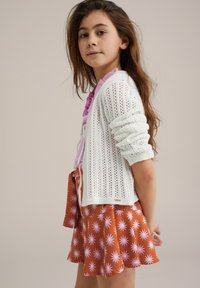 Fehér kötött kardigán mintás dizájnnal, rózsaszín ruffled felső fölött viselve, és egy barna szoknyán, amelyet rózsaszín margarétás motívumok díszítenek.