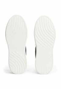 Suole di scarpe da ginnastica nere con fondi in gomma bianca, caratterizzate da motivi di trazione curvi e una leggera embossatura del logo su ciascuna suola.