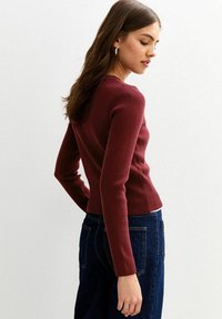 Bordeaux geribbelde trui met lange mouwen en aansluitend ontwerp. Gecombineerd met donkere spijkerbroek, vanuit een achteraanzicht. Subtle textuurdetails aanwezig.