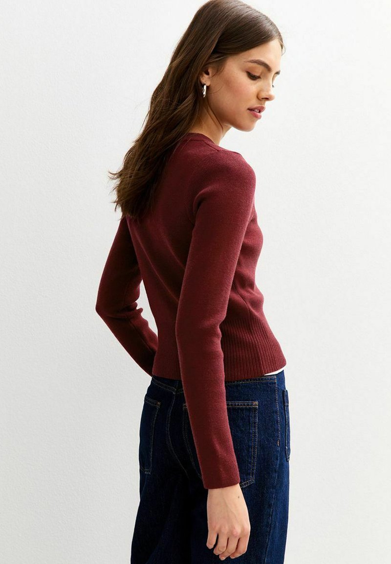 Burgunderfarbener, gerippter Pullover mit langen Ärmeln und figurbetontem Schnitt. Kombiniert mit dunklen Jeans, zeigt eine Rückansicht. Subtile Texturdetails sind vorhanden.