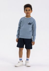 Blauwe katoenen sweater met een voorzak en geborduurde tekst, gecombineerd met marineblauwe cargoshorts, witte sneakers en gestreepte sokken.
