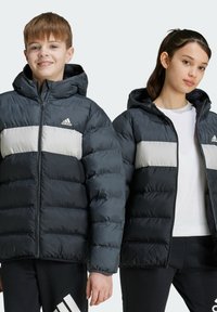 Chaquetas tipo puffer en gris oscuro y gris claro con capucha, cierre de cremallera y textura acanalada; logo en la parte superior izquierda; combinadas con pantalones más oscuros.