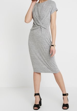Femme portant une robe grise, longueur genoux, manches courtes avec un détail torsadé à l'avant et des sandales noires à talons hauts avec des brides à la cheville.
