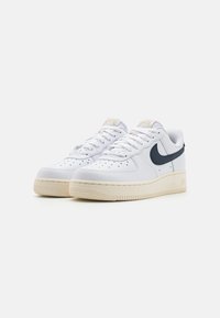 Een paar witte low-top Nike Air Force 1 sneakers met zwarte swoosh-logo's, crèmekleurige zolen en witte veters, weergegeven op een witte achtergrond.