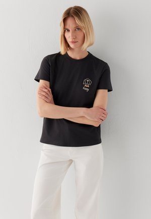 Frau mit kurzen blonden Haaren, die ein schwarzes T-Shirt mit einem kleinen Hundegesicht und dem Schriftzug „happy“ trägt, kombiniert mit weißen Hosen, steht vor einer weißen Wand.