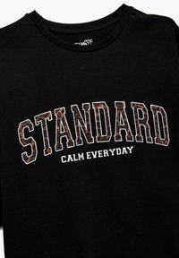 Camiseta negra de manga corta con la palabra "STANDARD" en estampado de leopardo marrón y "CALM EVERYDAY" en blanco, con cuello redondo, de material de algodón.
