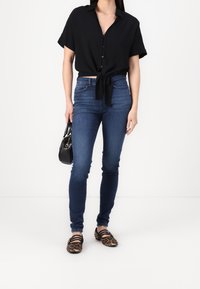 Camisa negra de botones con un lazo en la cintura, combinada con jeans ajustados azules y bailarinas negras con estampado de leopardo. Bolso negro en la mano.