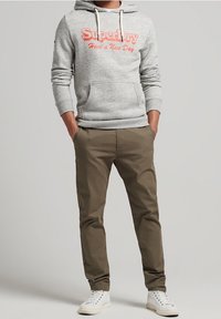Felpa grigia con cappuccio e logo "SuperDry" arancione, abbinata a pantaloni kaki e sneakers bianche. Presenta cappuccio con coulisse e tasca frontale.