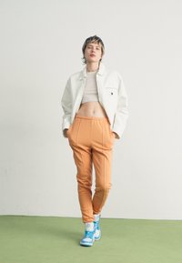 Fehér rövidített dzseki, bézs cropped top, narancssárga sportnadrág oldalsó varrással, és kék tornacipő. A modell zöld padlón áll, világos háttér előtt.