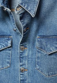 Veste en denim à l'avant avec boutons-pression métalliques, deux poches à rabat sur la poitrine, et coutures visibles sur un tissu bleu texturé.