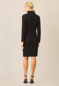 Schwarzes geripptes Strickkleid mit langen Ärmeln und hohem Kragen, das bis zur Mitte des Oberschenkels reicht. Kombiniert mit schwarzen Strappy-High-Heels.