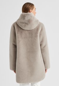 Frau in einem hellgrauen, weichen Kapuzenmantel, von hinten gezeigt, vor einem schlichten weißen Hintergrund stehend.