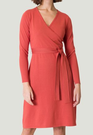 Femme portant une robe portefeuille corail à manches longues et longueur au genou, avec une ceinture nouée à la taille, sur un fond uni.