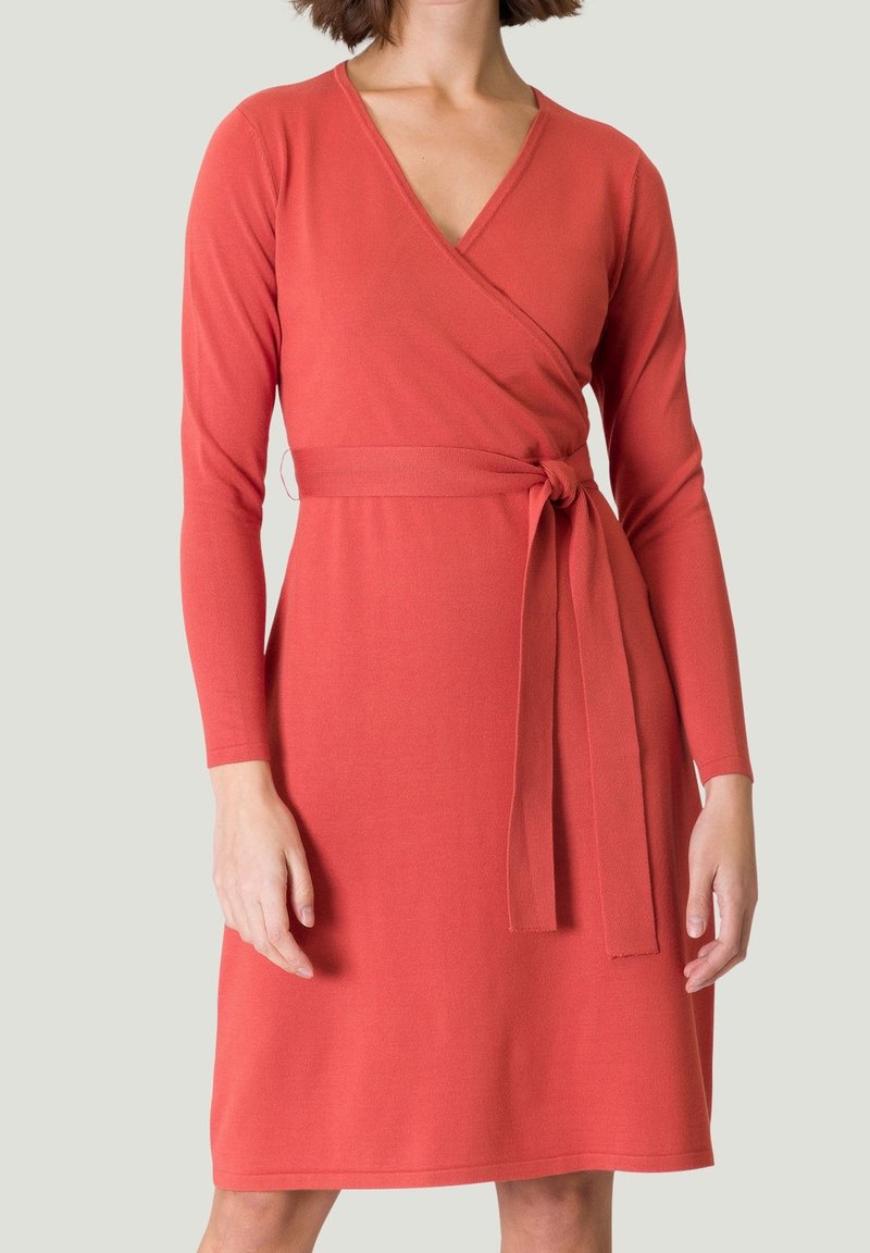 Femme portant une robe portefeuille corail à manches longues et longueur au genou, avec une ceinture nouée à la taille, sur un fond uni.