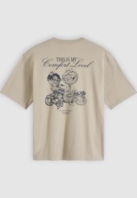 Smėlio spalvos medvilniniai marškinėliai su spaudiniu ant nugaros, vaizduojančiu grafines desertų iliustracijas ir tamsiu rašalu užrašytu tekstu „THIS IS MY Comfort Level“.