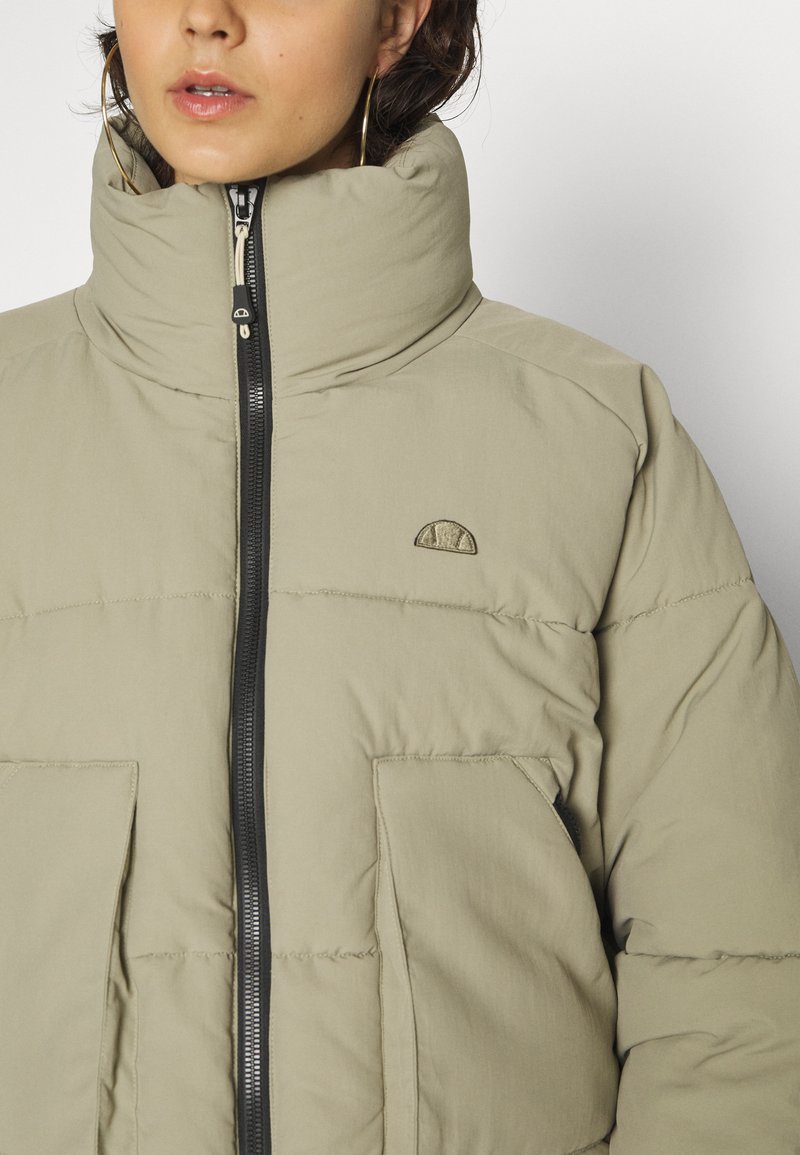 Jasno beżowa kurtka puchowa z wysokim kołnierzem, z przodu na zamek błyskawiczny oraz dużymi kieszeniami. Zawiera małe logo na klatce piersiowej oraz teksturowaną powierzchnię.