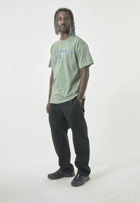 Green cotton t-shirt with blue text and logo, paired with black pants and black sneakers with colorful accents. Casual fit and relaxed design.
