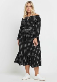 Svart off-shoulder-klänning med vitt geometriskt mönster, volangkjol och elastisk midja. Matchad med vita sneakers för en avslappnad look.