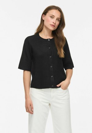 Femme portant un cardigan noir à manches courtes boutonné et un pantalon blanc, debout avec une main dans sa poche devant un fond uni.