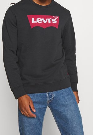 Man draagt een zwarte Levi’s trui met rood logo en blauwe jeans, staand tegen een lichte, effen achtergrond.