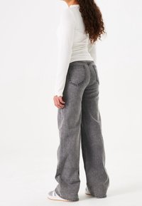 Vaqueros de pierna ancha gris con un acabado texturizado, que presentan costuras visibles y bolsillos de parche, combinados con una camiseta ajustada de manga larga blanca.