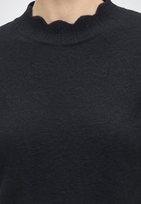 Gros plan d'un pull en maille noir avec un col montant froncé porté par une personne.