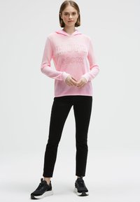 Junge Frau steht mit gefalteten Händen, trägt ein pinkes Mesh-Hoodie mit dem Text "HOPE soccx", schwarze Hosen und schwarze Sneakers.