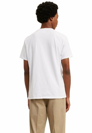 Mann mit lockigem Haar, der ein schlichtes weißes Kurzarm-T-Shirt und beige Hosen trägt, steht mit dem Rücken zu einem weißen Hintergrund.