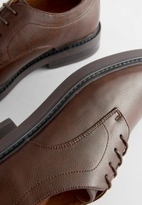 Chaussures habillées en cuir marron à lacets avec des coutures détaillées et des semelles noires sur un fond clair.