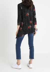 Tunique noire avec motifs floraux en rouge et rose, manches trois-quarts, ourlet asymétrique, associée à un jean skinny bleu et des baskets blanches.